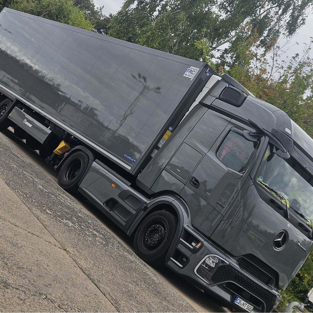 Grauer Mercedes-Lkw mit Plane auf einem Parkplatz, im Hintergrund Bäume.