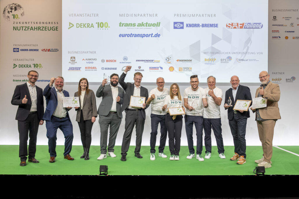 Gruppe von zehn Personen auf Bühne mit Urkunden und Daumen hoch vor Werbewand.