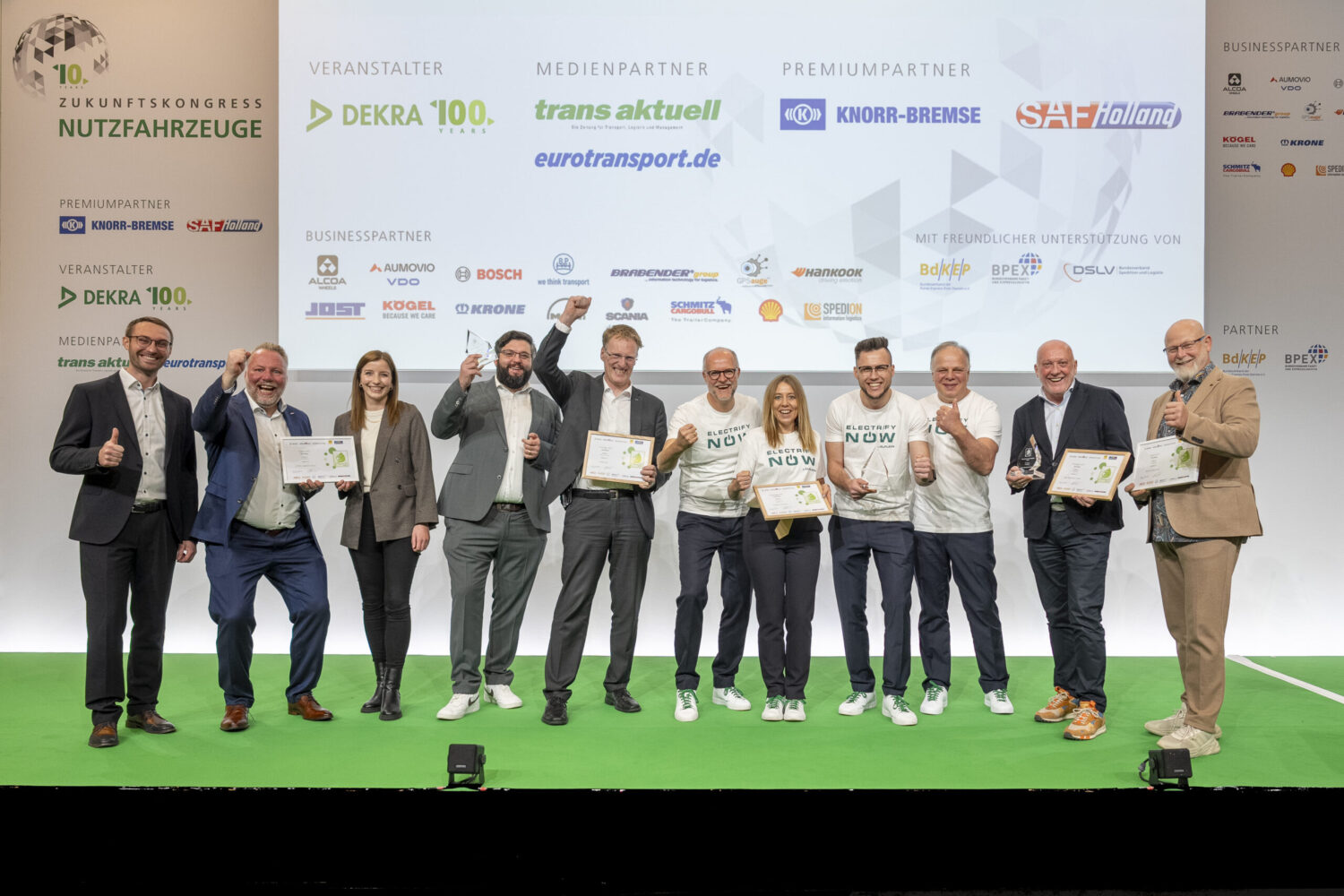 Gruppe von zehn Personen auf Bühne mit Urkunden und Daumen hoch vor Werbewand.